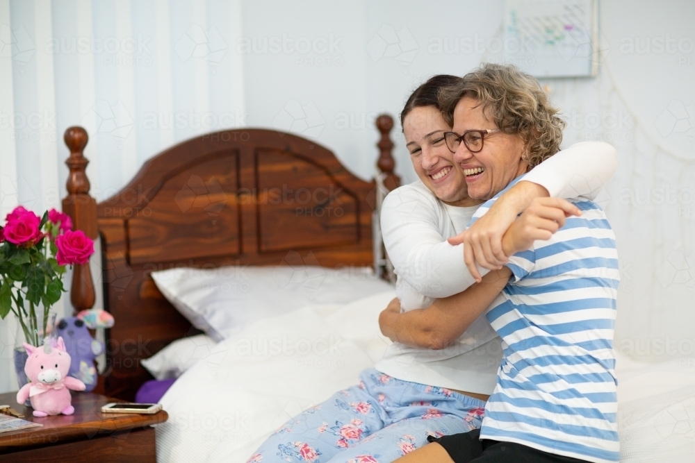 Mother and teen daughter sitting on bed and laughing with arms around each other - Australian Stock Image
