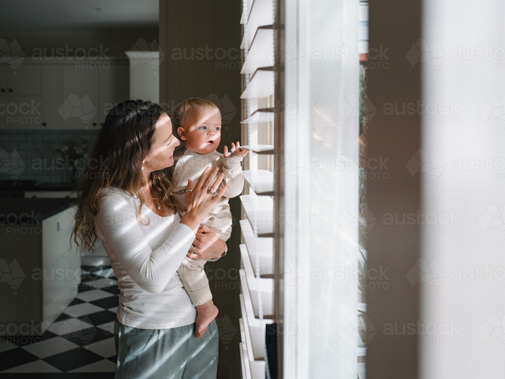 Mother carrying baby peeking through window blinds - Australian Stock Image