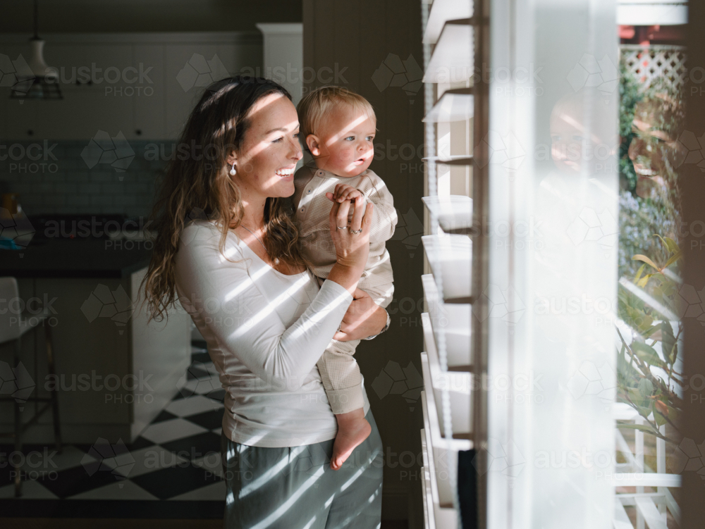 Mother carrying baby peeking through window blinds - Australian Stock Image