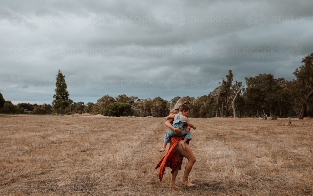 Mother carrying her son through paddock with overcast clouds - Australian Stock Image