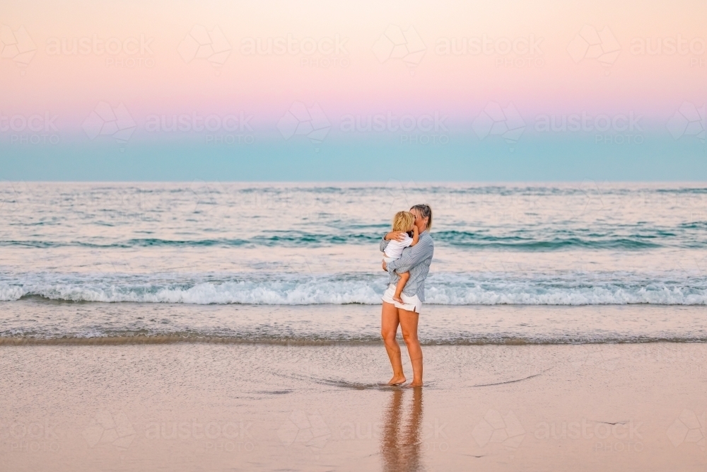Mother carrying son on the beach in pink twilight light. Sunset on the Gold Coast. - Australian Stock Image