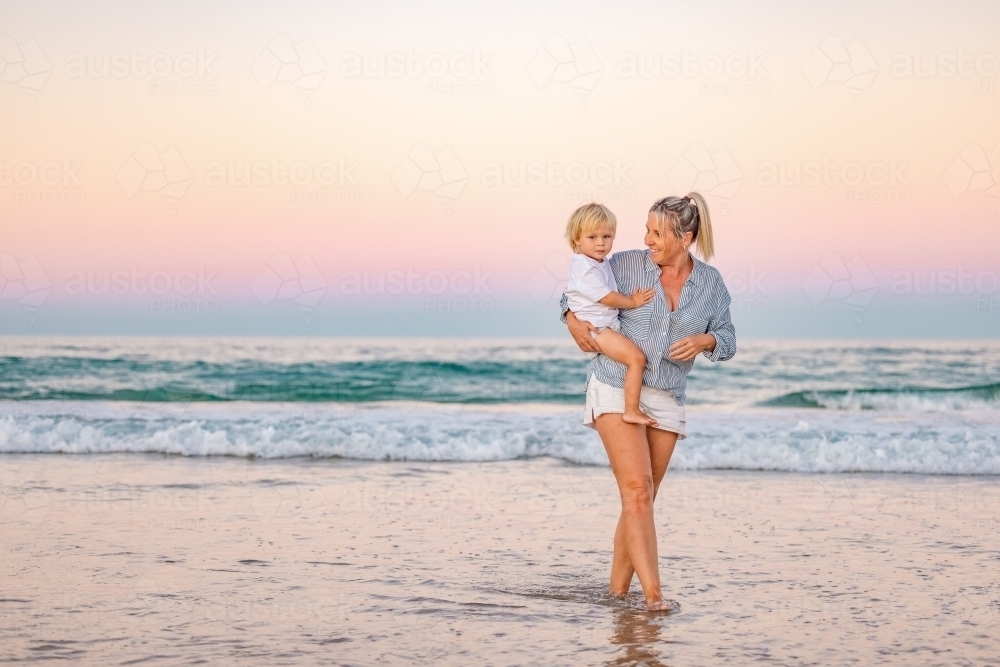 Mother carrying son on the beach in pink twilight light. Sunset on the Gold Coast. - Australian Stock Image