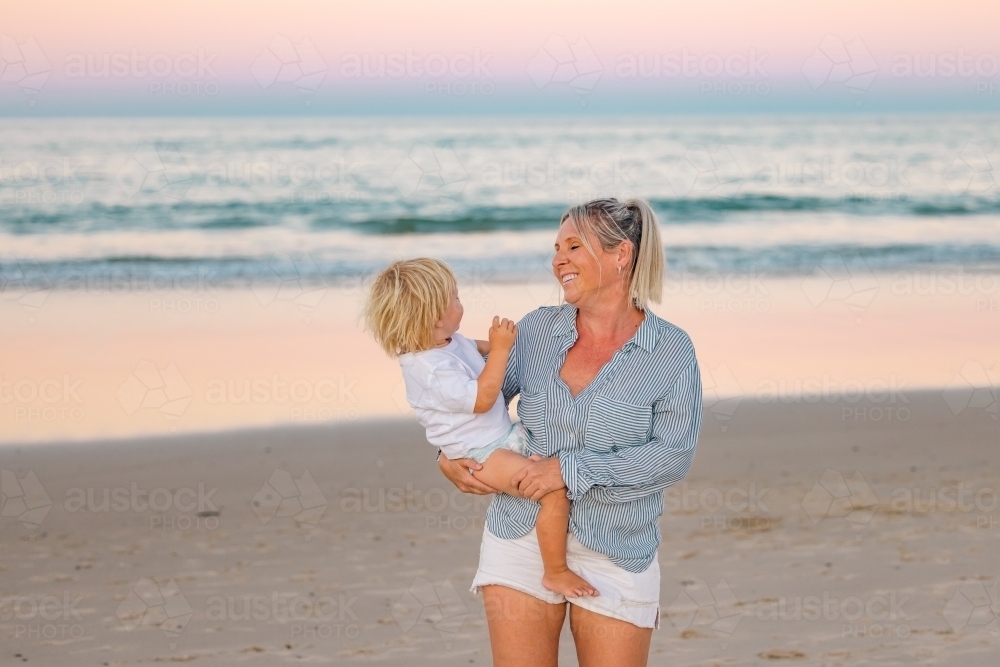 Mother carrying son on the beach in pink twilight light. Sunset on the Gold Coast. - Australian Stock Image