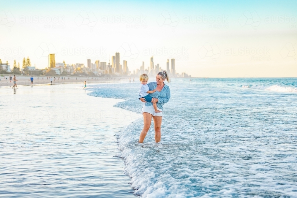 Mother carrying son on the beach with Gold Coast city skyline in background - Australian Stock Image