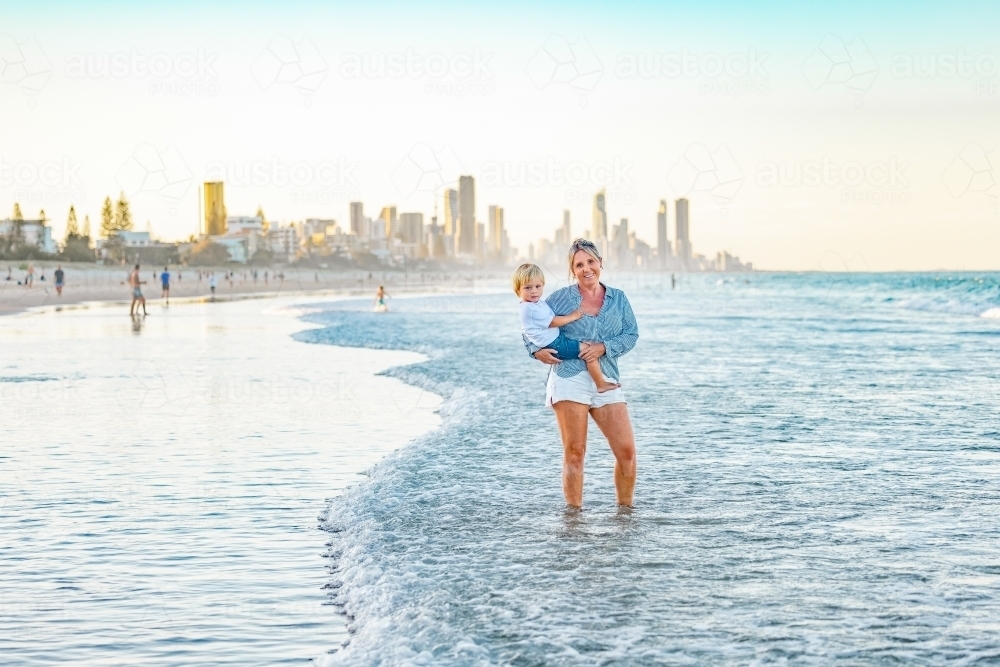 Mother carrying son on the beach with Gold Coast city skyline in background - Australian Stock Image