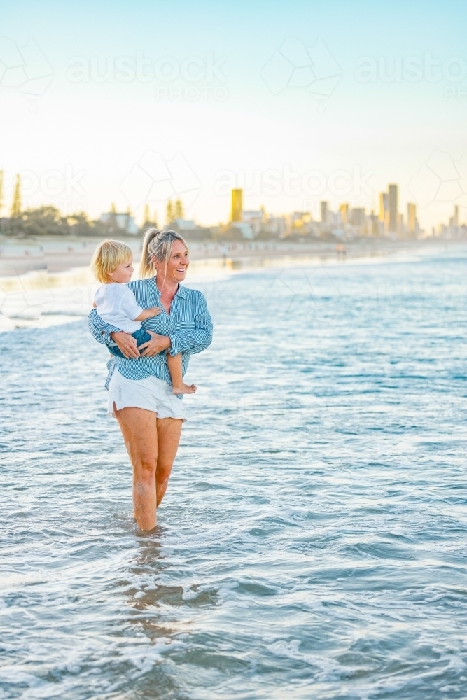 Mother carrying son on the beach with Gold Coast city skyline in background - Australian Stock Image