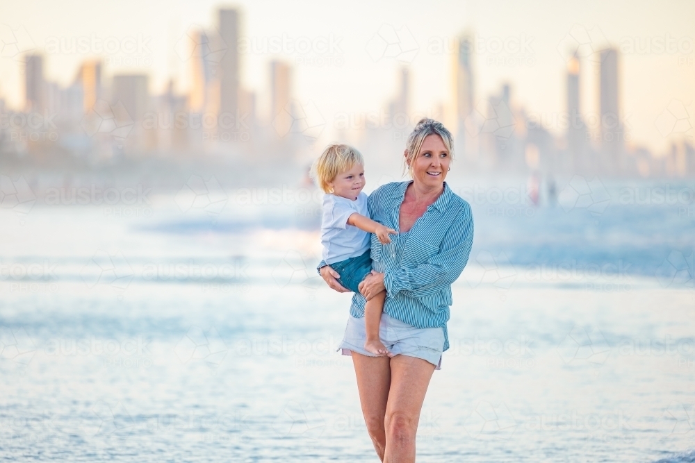 Mother carrying son on the beach with Gold Coast city skyline in background - Australian Stock Image