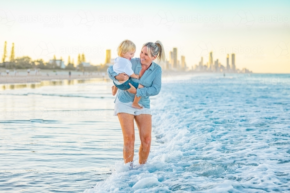 Mother carrying son on the beach with Gold Coast city skyline in background - Australian Stock Image