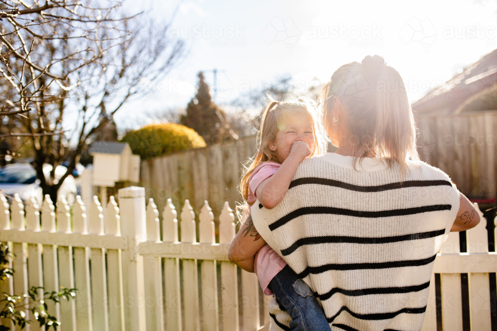 Mother carrying toddler daughter on her side beside picket fence - Australian Stock Image