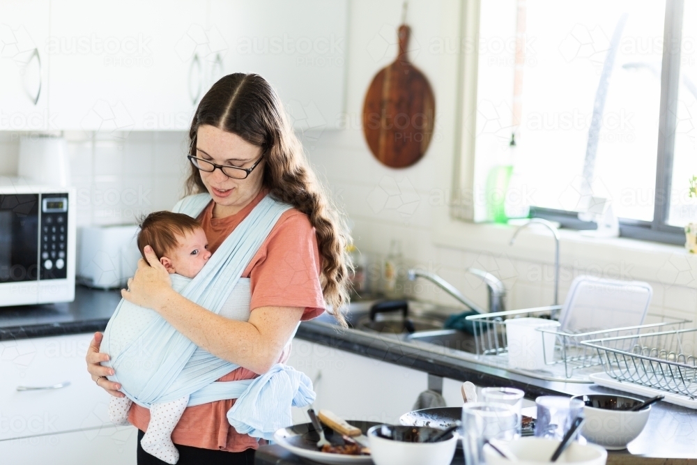 Mother cleaning up messy kitchen piled with dirty dishes while carrying newborn baby in wrap - Australian Stock Image