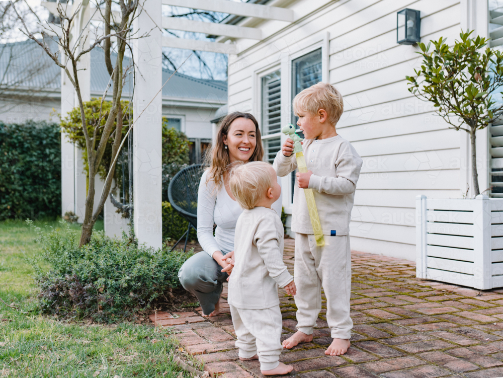Mother crouching on brick patio playing bubbles with two sons - Australian Stock Image