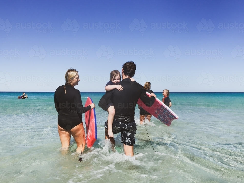 Mother, father and toddler in wetsuits, walking out in the ocean holding boogie boards - Australian Stock Image