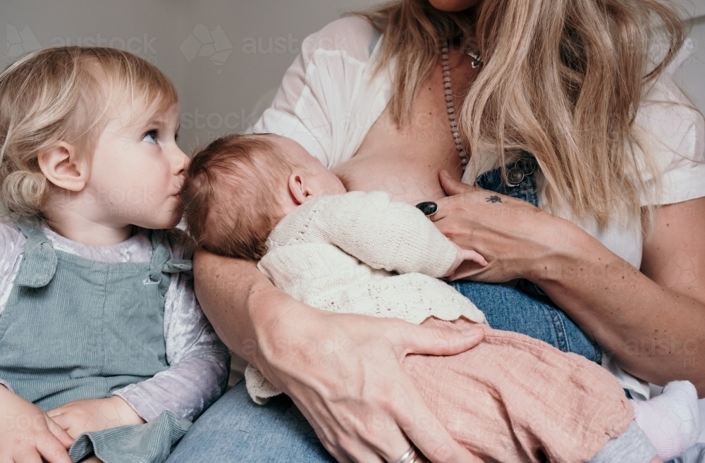 Mother feeds baby with toddler kissing her new sister. - Australian Stock Image