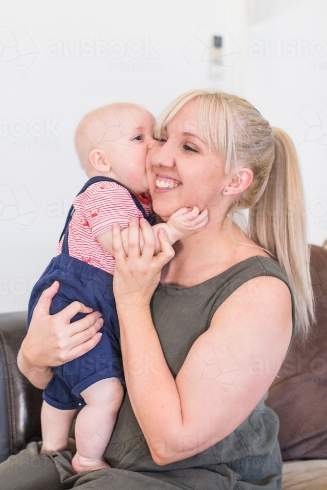 Mother holding baby boy smiling while he kisses her cheek - Australian Stock Image