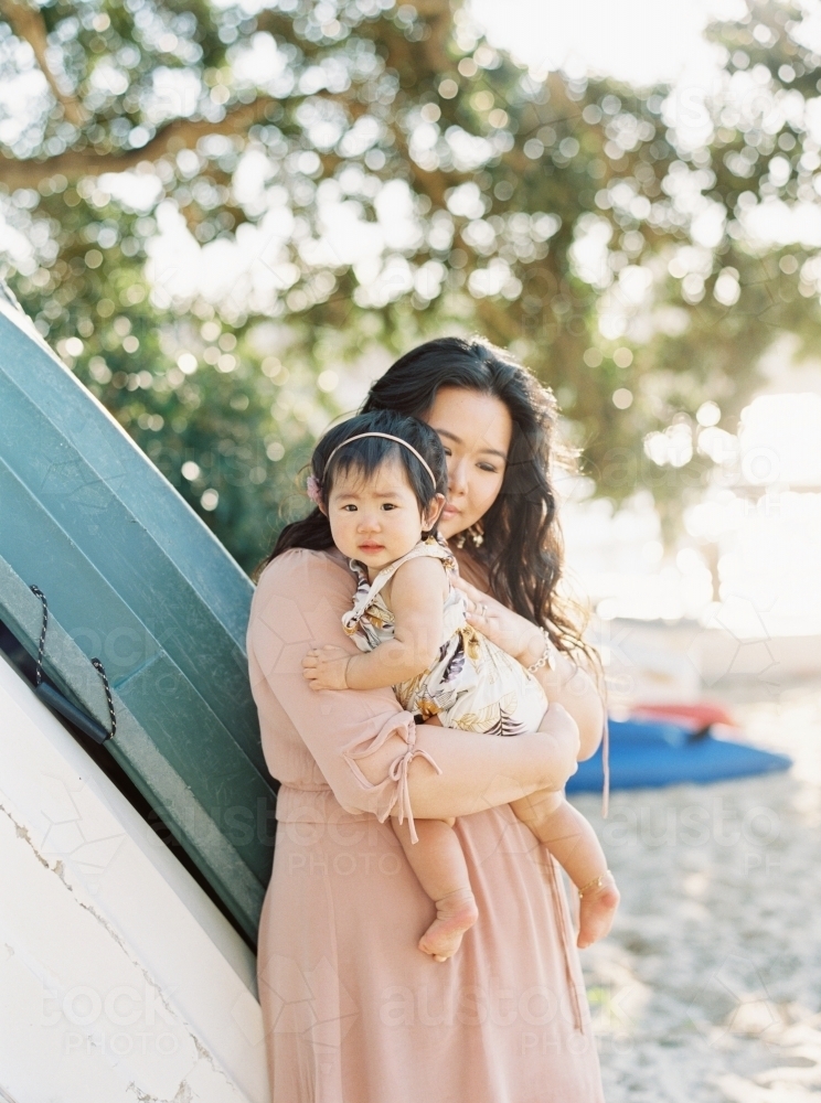 Mother holding baby girl at the beach, leaning against some boats - Australian Stock Image