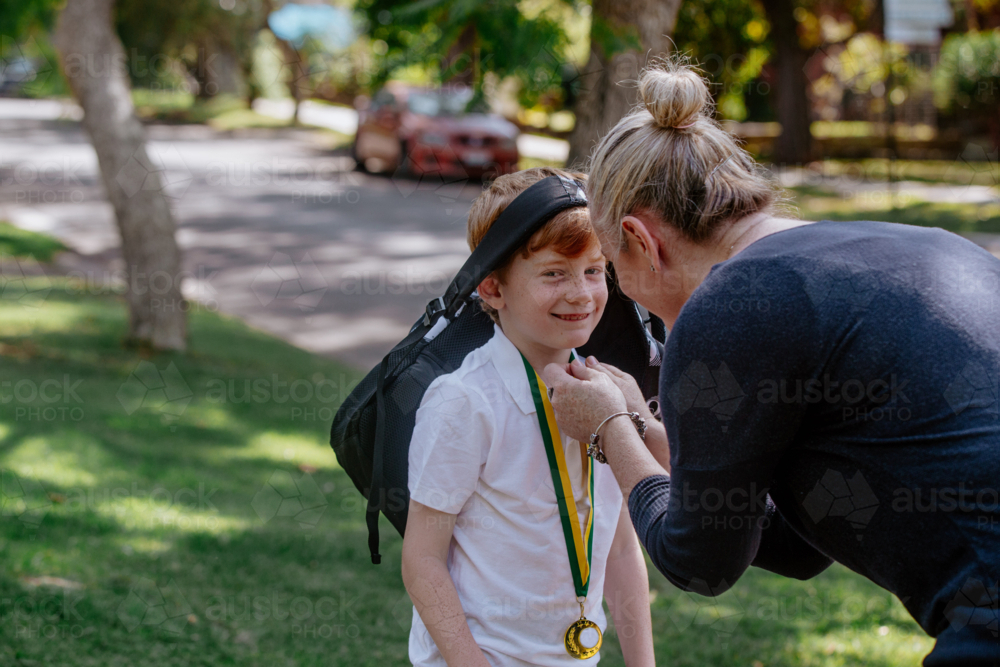 Mother holding cheeks of a happy young schoolboy with bag strap on the head - Australian Stock Image