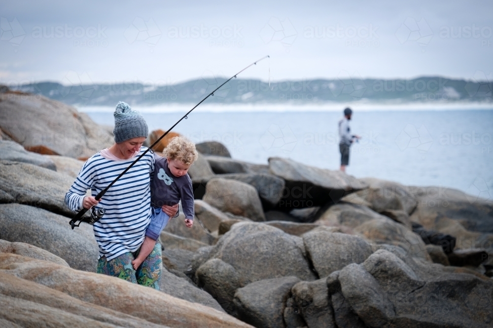 Mother holding daughter walking through rocks on beach with fisherman in background coastal backdrop - Australian Stock Image