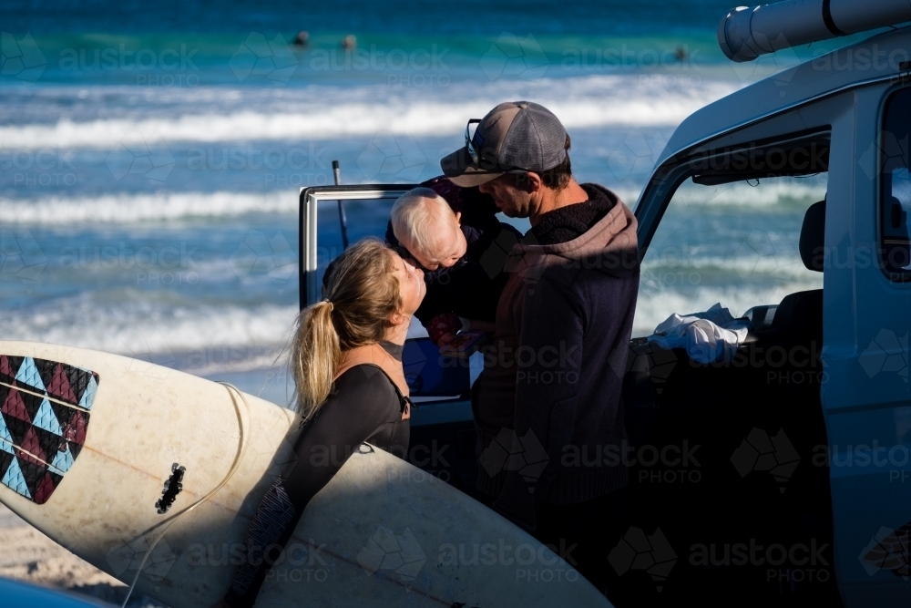 Mother holding surfboard kissing goodbye child in fathers arms beside 4WD at beach - Australian Stock Image