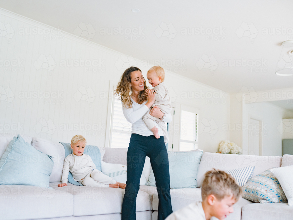 Mother holding toddler baby with young boys playing around her in the living room - Australian Stock Image
