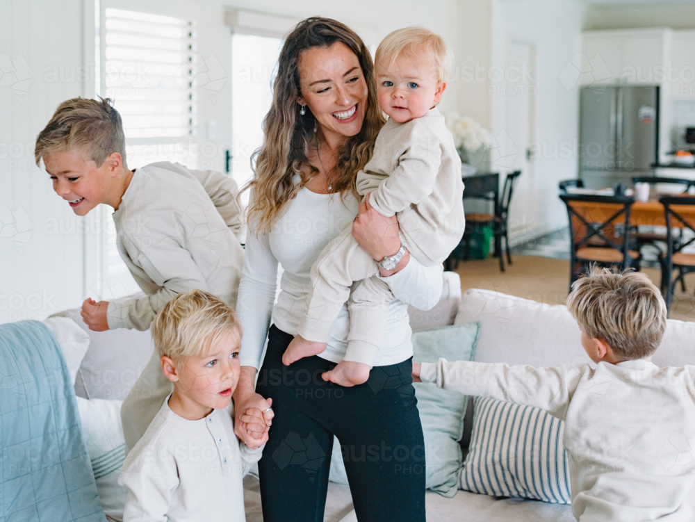 Mother holding toddler baby with young boys playing around her in the living room - Australian Stock Image