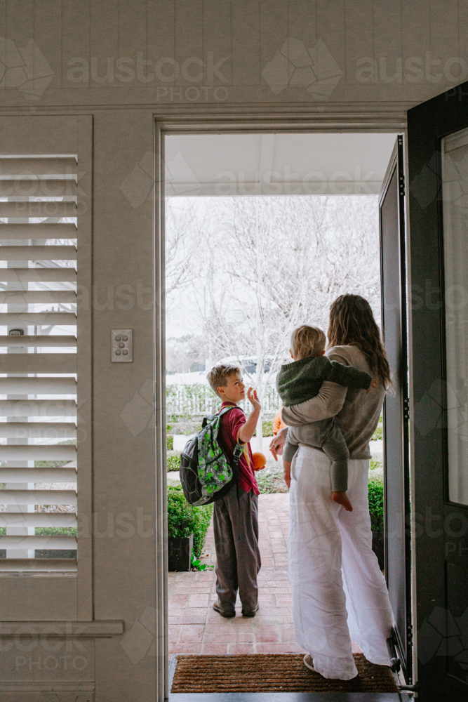 Mother holding toddler son in her hip saying goodbye to older son with backpack out the doorway - Australian Stock Image