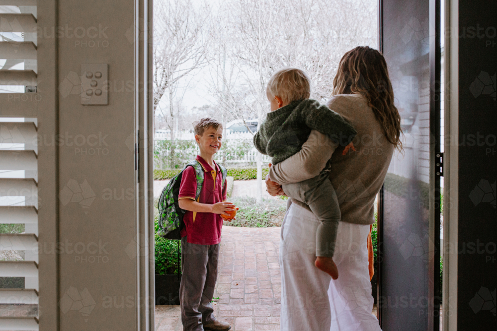 Mother holding toddler son in her hip saying goodbye to older son with backpack out the doorway - Australian Stock Image