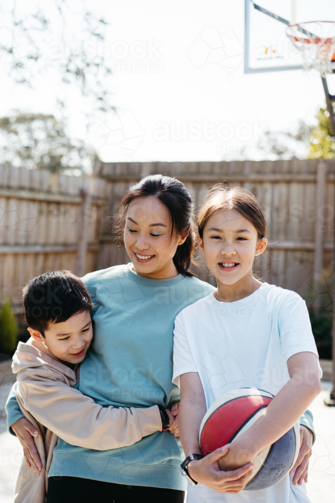 Mother hugs her two young children outside in their backyard. - Australian Stock Image