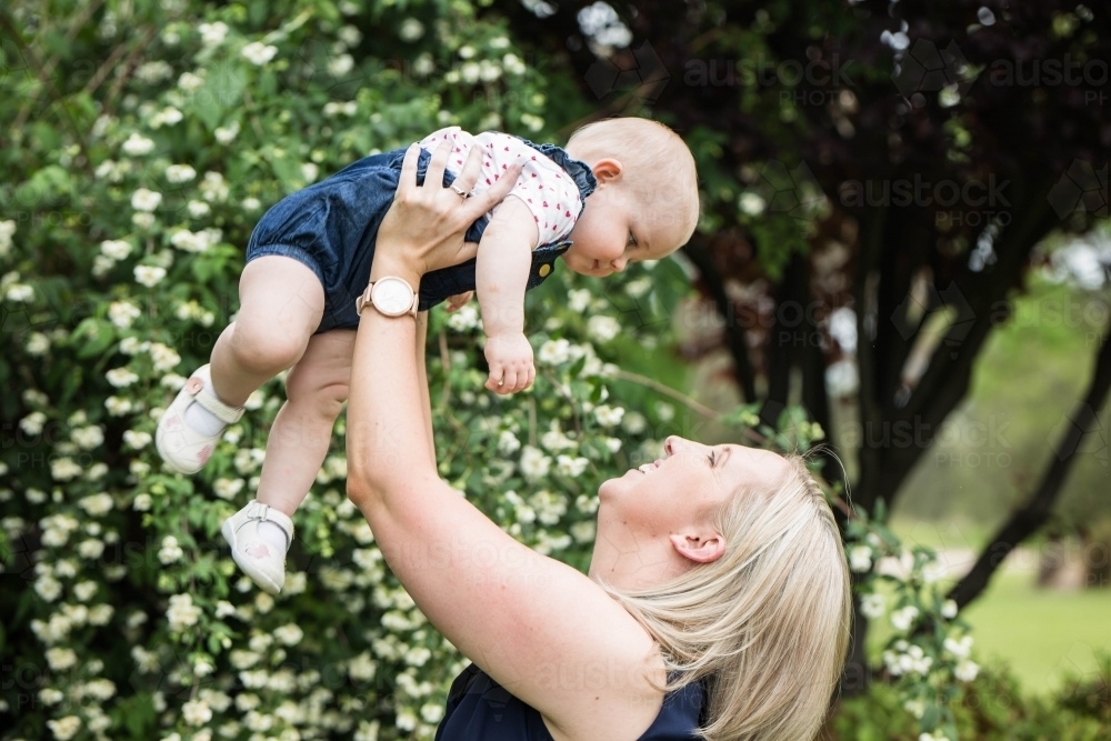 Mother lifting baby up in air with flowers in background - Australian Stock Image