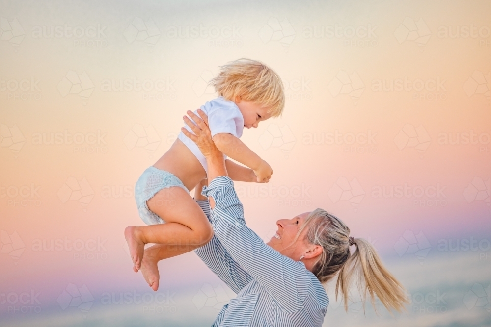 Mother lifting toddler son in the air on the beach with pink sunset sky - Australian Stock Image