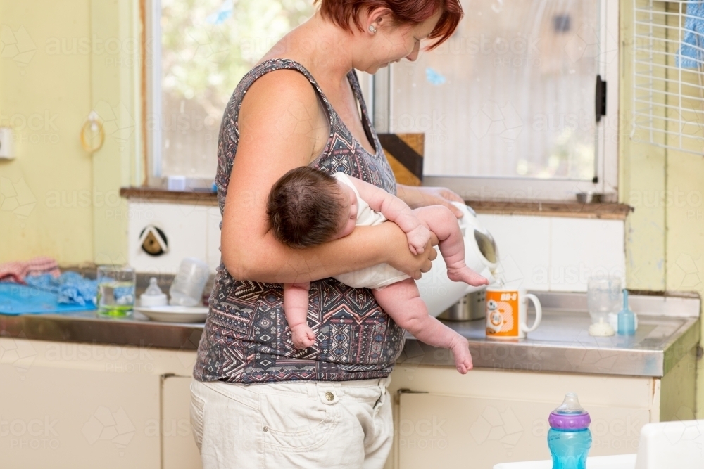 Mother making cup of tea while holding newborn baby - Australian Stock Image