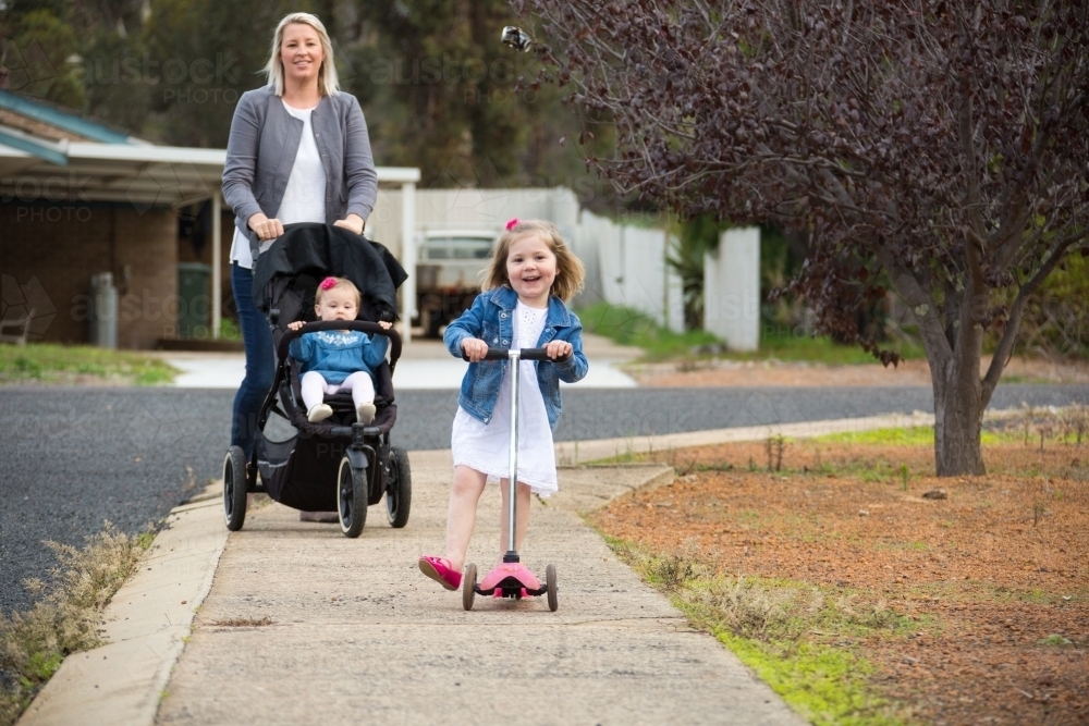 Mother pushing baby in pram and young daughter on scooter - Australian Stock Image