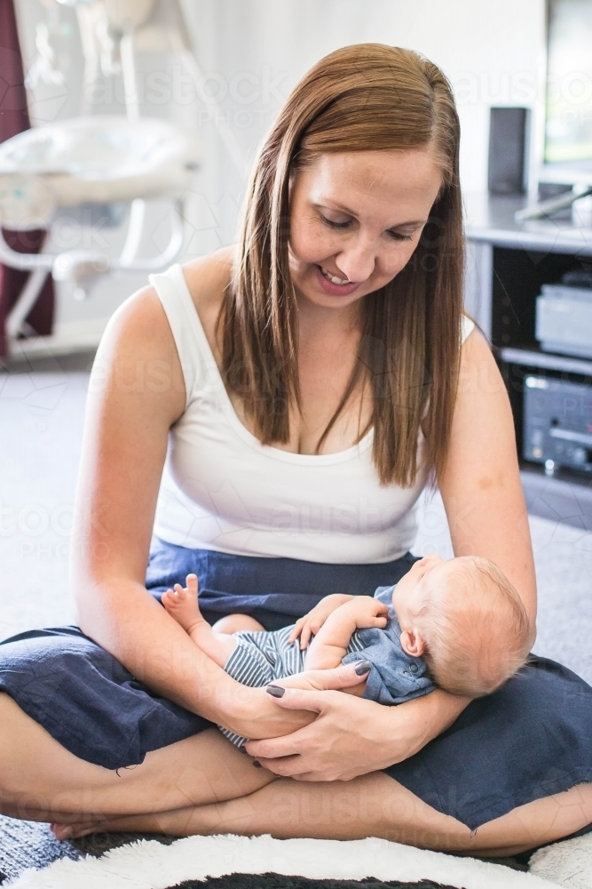 Mother sitting on floor at home smiling down at newborn baby in arms - Australian Stock Image
