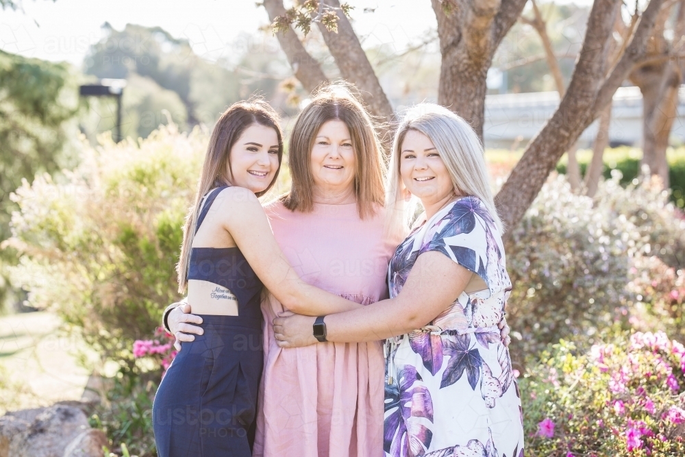 Mother standing with arms around two daughters hugging and smiling - Australian Stock Image