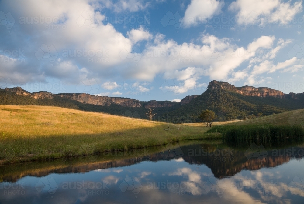 Mountain escarpment, sunlit grass and clouds reflected in farm dam - Australian Stock Image