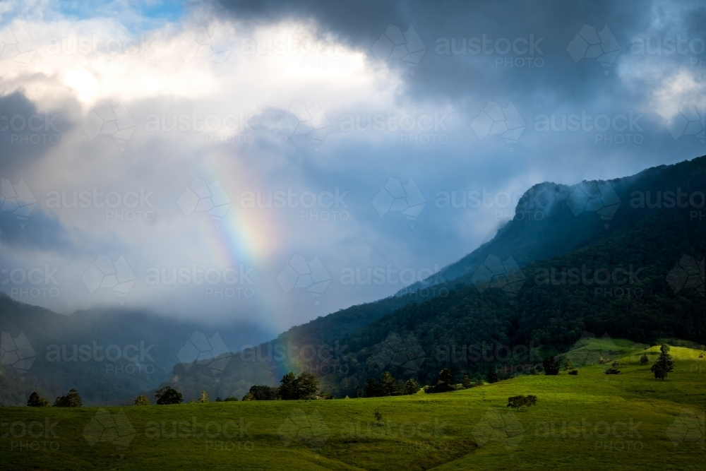 Mountain range covered by clouds with sunlight and rainbow - Australian Stock Image
