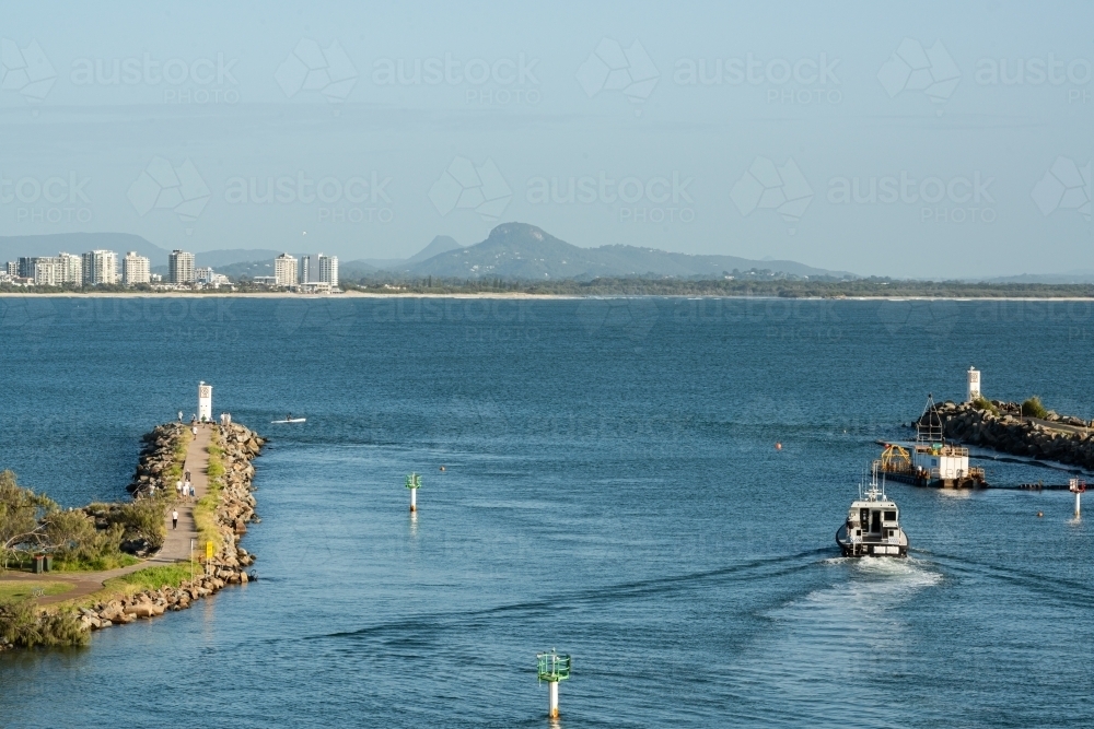 Mouth of a river with people walking on groyne, boat motoring out, high rise buildings and mountains - Australian Stock Image