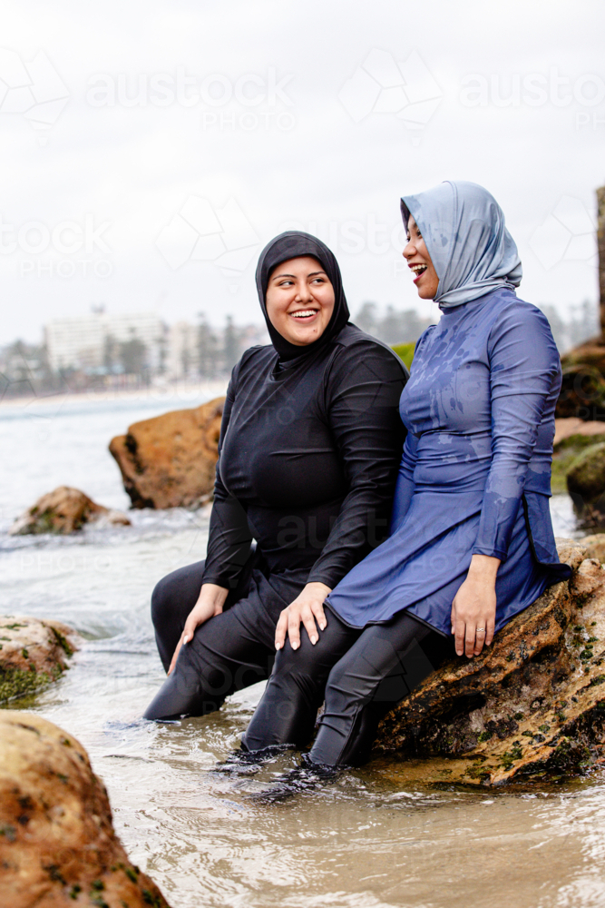 Multiracial friends wearing headscarves sitting on the large shoreline rocks - Australian Stock Image