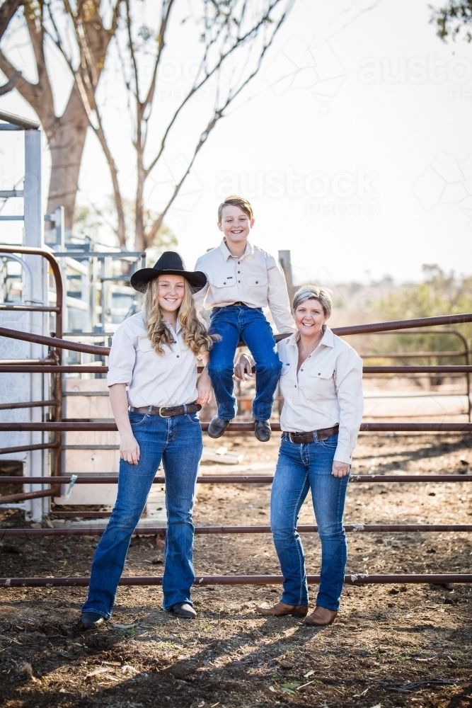 Mum and daughter standing with son sitting on gate in yards on farm in drought - Australian Stock Image