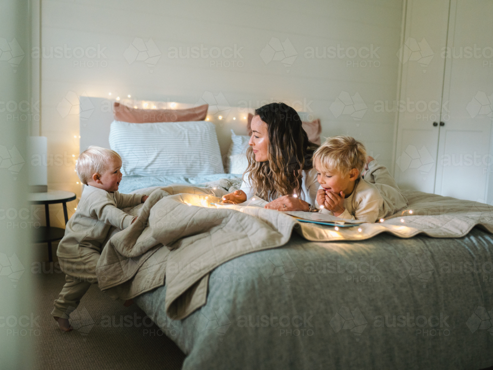 Mum and son lying on their belly and toddler baby climbing on the bed on the bed with string lights - Australian Stock Image