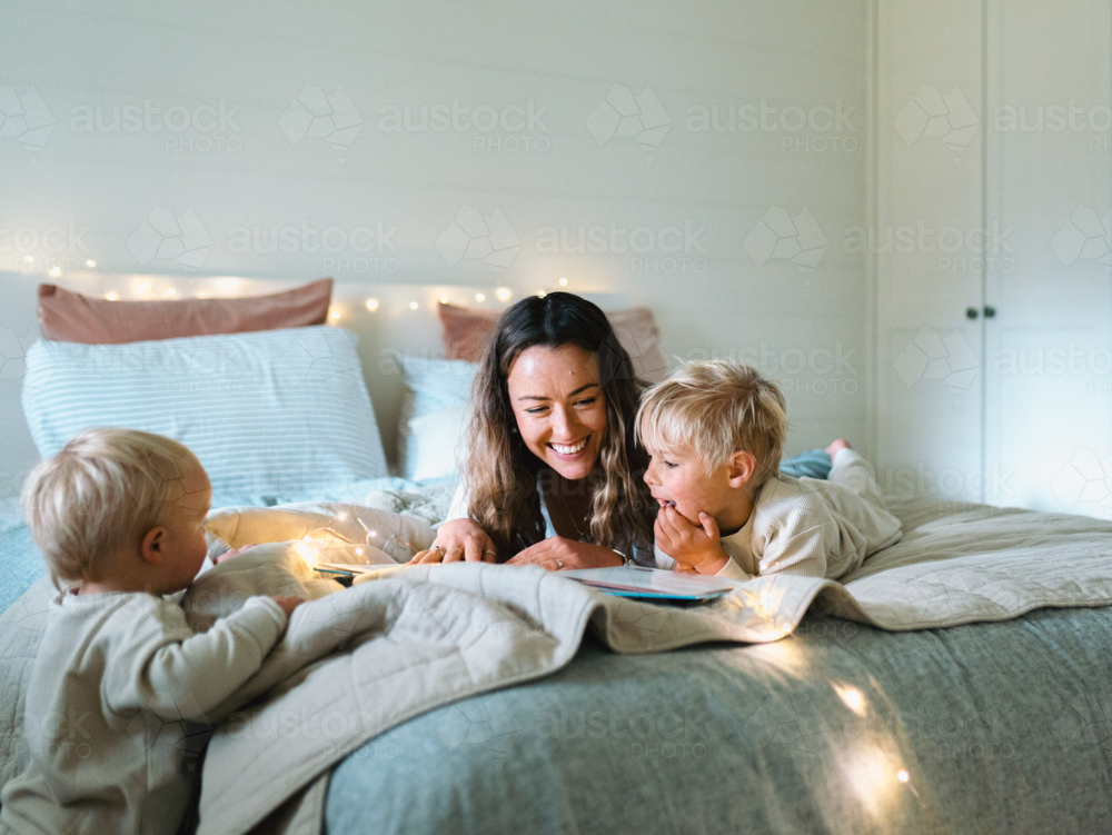 Mum and son lying on their belly on the bed with string lights and toddler standing on bedside - Australian Stock Image
