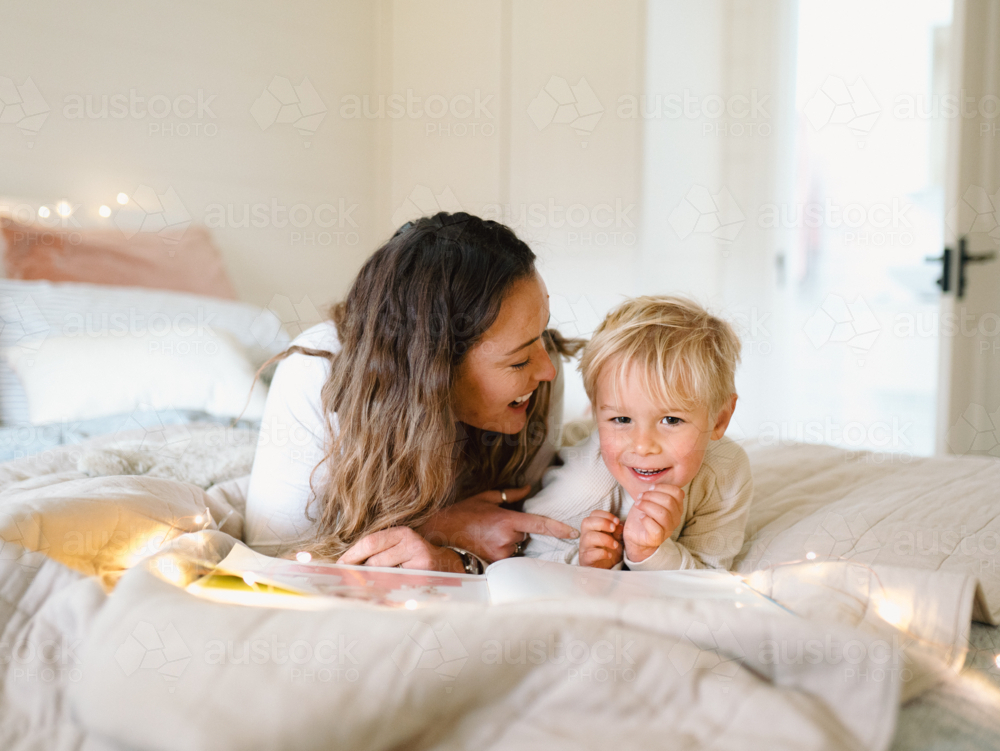 Mum and toddler son lying on their belly on the bed with string lights reading a book - Australian Stock Image