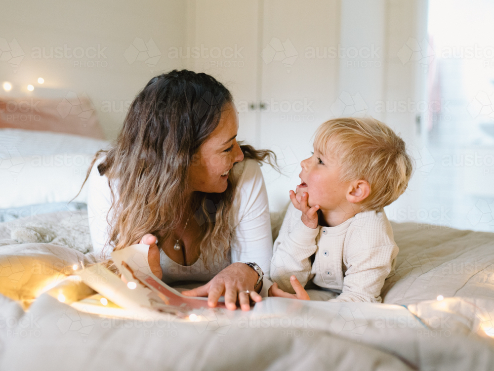 Mum and toddler son lying on their belly on the bed with string lights reading a book - Australian Stock Image