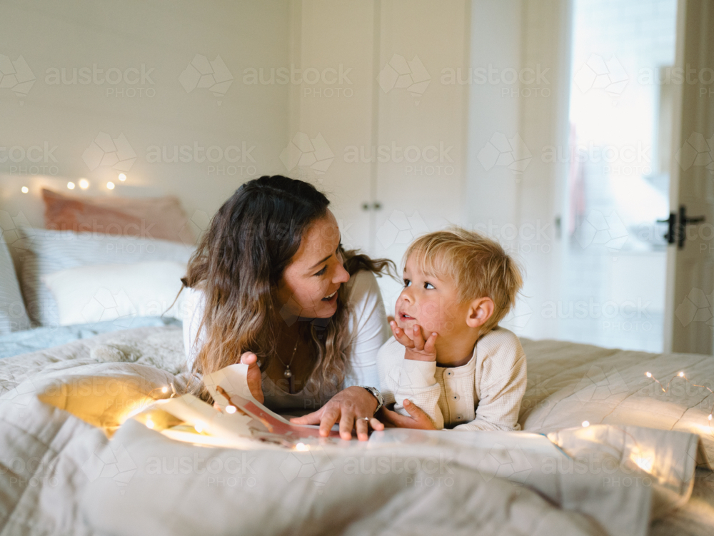 Mum and toddler son lying on their belly on the bed with string lights reading a book - Australian Stock Image