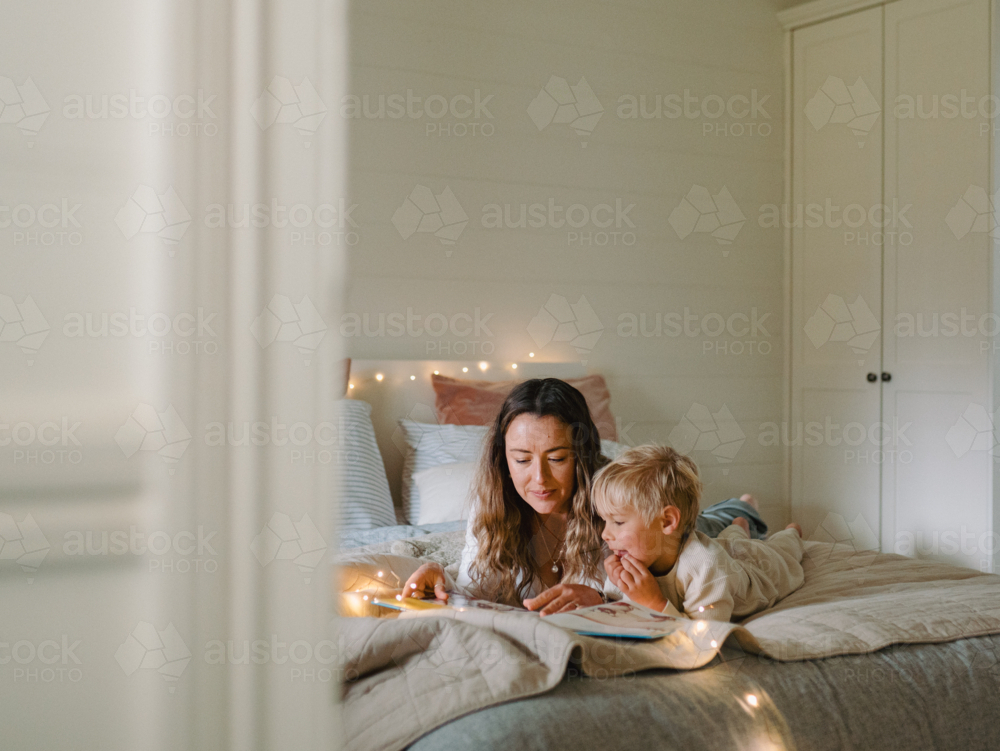 Mum and toddler son lying on their belly on the bed with string lights reading a book - Australian Stock Image