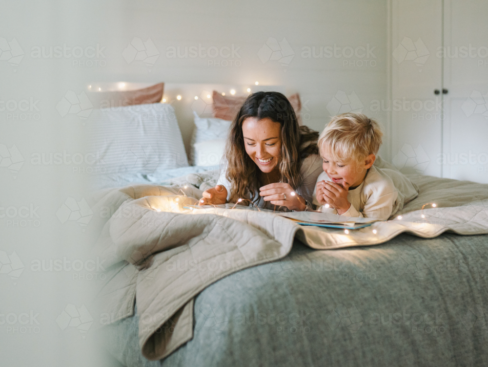Mum and toddler son lying on their belly on the bed with string lights reading a book - Australian Stock Image