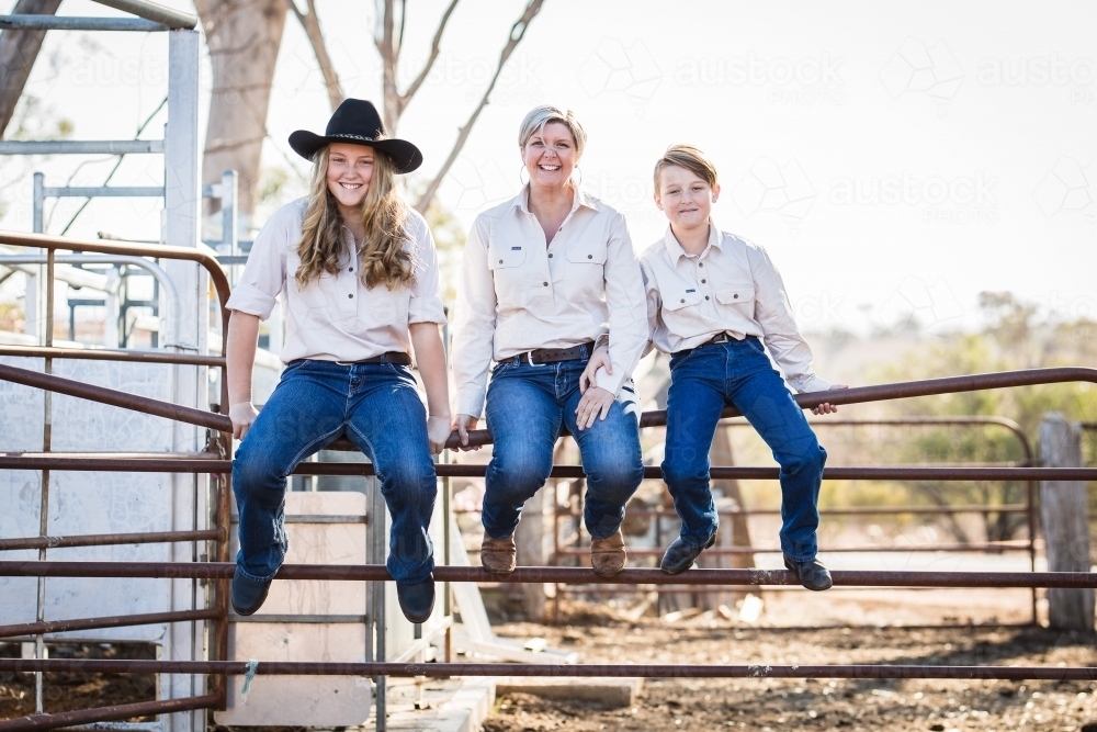 Mum and two children sitting on stock yards gate smiling on farm in drought - Australian Stock Image