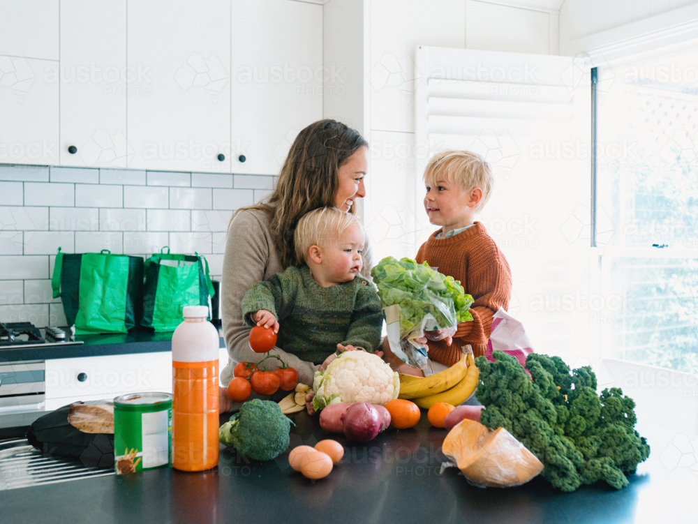 mum and young boys sitting on the kitchen countertop by fresh fruits and vegetables from the shops - Australian Stock Image