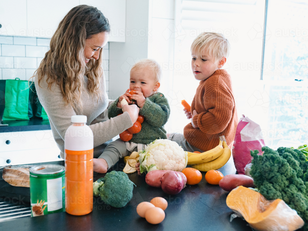 Mum holding Baby boy taking a bite on a bag of tomato while older brother looking at him - Australian Stock Image