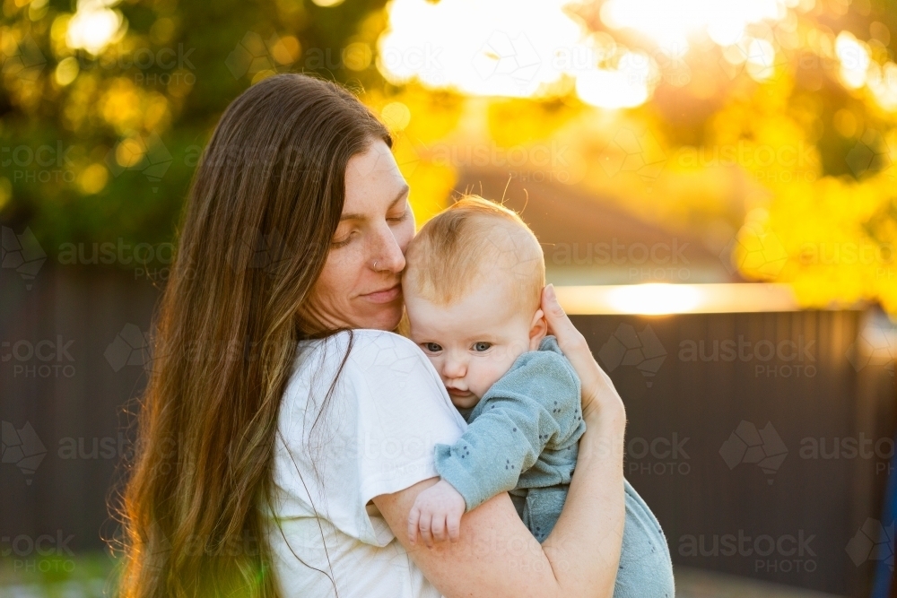 mum holding four month old baby girl in arms outside in golden bokeh sunset light in backyard - Australian Stock Image