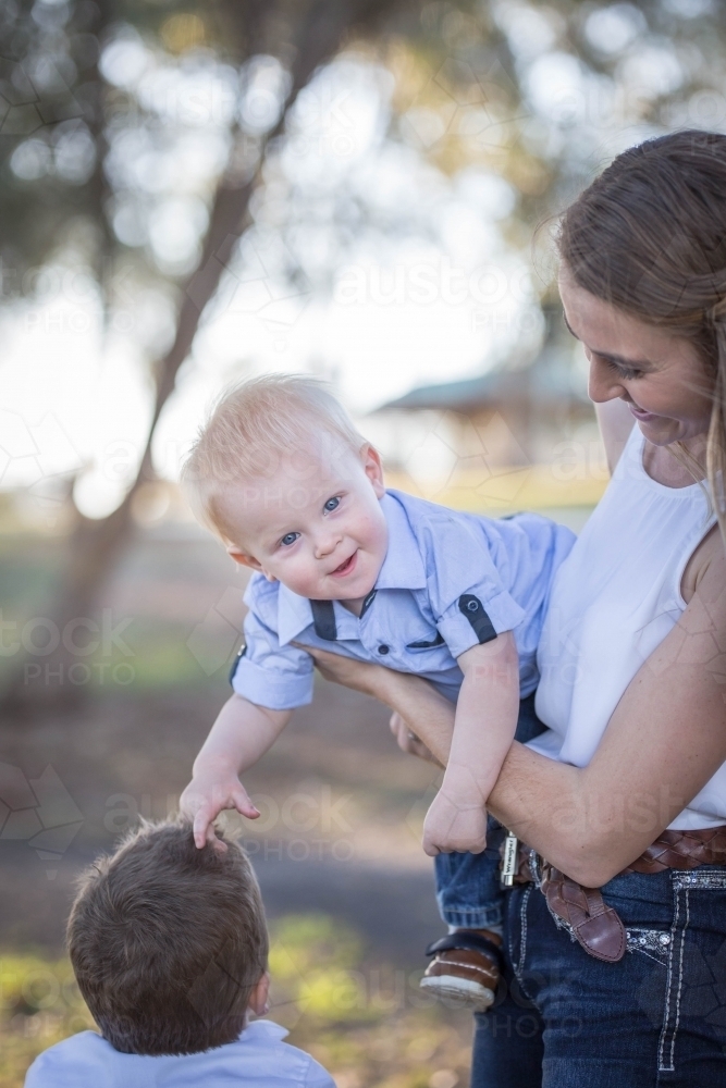 Mum holding laughing baby boy on hip while he plays with brother's hair - Australian Stock Image